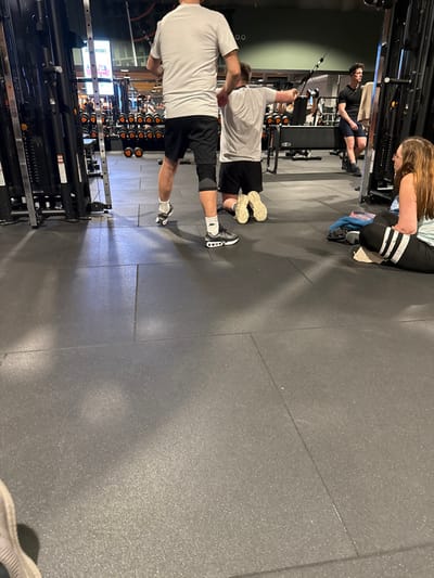 Two men are exercising at the gym, pulling a cable machine. The man in the foreground has a white t-shirt, black shorts and white sneakers, and his stance is wide. A younger man behind him wears similar attire. A woman is sitting on a bench nearby, wearing workout leggings with white stripes. In the background are rows of weight equipment and another man is sitting on a bench. The gym has a gray, tiled floor.