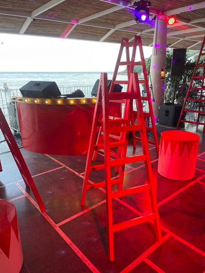 This image captures an outdoor event space in Menderes, Turkey, featuring a vibrant, circus-themed setup overlooking the Aegean Sea under an overcast sky. The scene is bathed in artificial red and purple lighting.

In the foreground and midground, several red A-frame ladders are prominently positioned on a dark floor that is demarcated by white tape forming a grid pattern. Two red cylindrical stools, each adorned with a white pointed pattern along the top edge, are visible; one is on the far right and another is partially shown on the far left.

To the left, a curved red DJ booth or bar with a wooden countertop is present, featuring a row of exposed bulbs. On the counter, two black speakers flank what appears to be DJ equipment, partially covered by a black fabric. Behind the booth, a white metal railing separates the venue from the sea, where a small white boat is visible.

On the right side of the midground, a substantial white, textured column supports the overhead structure. A lantern hangs from this column, which is also illuminated by purple light projections. A black speaker on a stand is positioned to the right of the column, with green foliage and distant land visible beyond.

The overhead canopy is constructed from a bamboo or straw covering, supported by white metal beams. Mounted on these beams are various stage lights, including a purple moving head fixture and a red LED par light, which cast the dominant colors onto the scene. No people are visibly present in the frame.