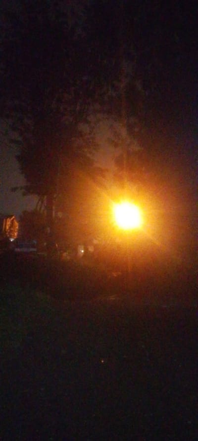 The image shows a dark, blurry scene at night. There is a bright, diffused light source in the center of the image. Trees and possibly buildings are silhouetted against the light. The foreground appears to be grassy. The location is Ndenderu ward, Kenya.