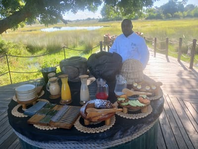 The scene depicts a breakfast buffet laid out on a round table outdoors, presumably in Nwanaeanoka, Botswana, based on the setting. A man wearing a white chef's coat with the "North Island Seychelles" logo stands behind the table. The table displays a variety of food items, including a stack of plates on a wooden stand, several pitchers containing various liquids (possibly juices and milk), a pound cake dusted with powdered sugar, sliced fruits and vegetables, and deviled eggs. The table itself is decorated with rope trim and is set on a wooden deck overlooking a marshy landscape with tall grasses and a body of water in the distance. Behind the man and the table are decorative wooden objects. The scene is bathed in sunlight, suggesting it is morning.