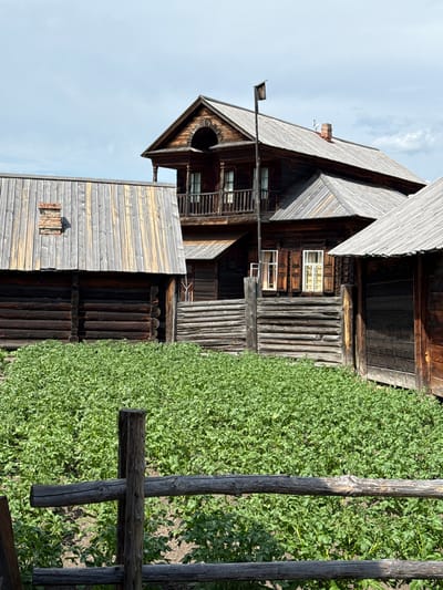 The image depicts a rural scene in Shushenskoye, Russia, likely part of an ethnographic museum or historical reconstruction. In the foreground, a rustic wooden fence runs horizontally across the frame, with a wooden post intersecting it. Behind the fence, a dense field of green plants, appearing to be potato plants, fills the lower half of the image.
Dominating the background are several wooden structures, characteristic of traditional Russian rural architecture. To the left, a long, low building with a sloped wooden roof and visible log construction is partially visible. A brick chimney extends from its roof. Adjacent to this, and slightly to the right, is a larger, multi-story wooden house. This house features dark, weathered logs, multiple windows with wooden shutters, and a balcony with a wooden railing on the upper level. A tall wooden pole with a small birdhouse-like structure at the top stands near the house. To the far right, another wooden structure, possibly an outbuilding or shed, is visible with a sloped wooden roof and dark wooden walls.
The sky above is partly cloudy with a light blue hue. The overall impression is one of a preserved historical rural environment.