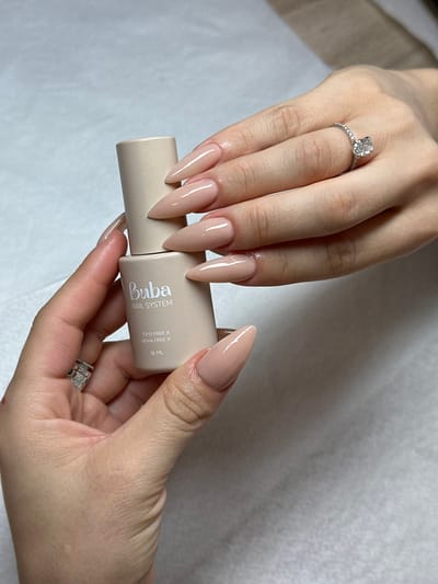 In Kafr Kasim, Israel, a close-up image captures two human hands on a light, textured surface. The hand on the lower left holds a cylindrical, light beige "Buba NAIL SYSTEM" bottle, labeled "TPD FREE X HEMA FREE X 16 ML." This hand wears a silver-toned, textured ring on its ring finger, and its fingernails are short. Positioned above and to the right, the second hand displays five long, pointed, almond-shaped nails, each uniformly coated with a glossy, light nude or beige polish. This hand features a silver-toned ring with an oval-cut clear gemstone, flanked by smaller stones, on its ring finger. The interaction suggests the product in the bottle is related to the nail application on the upper hand.