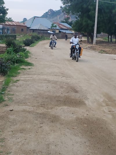 This media file depicts a scene in Ampan, Nigeria. The primary subjects are two motorcycles with riders. The first motorcycle, closer to the foreground, is ridden by a person wearing a white shirt and dark pants. They are looking ahead as they travel. Behind this motorcycle, another person is riding a second motorcycle, also appearing to be moving forward.

The setting is an unpaved, dusty road that curves to the right as it recedes into the distance. On the left side of the road, there are several buildings with corrugated metal roofs, characteristic of rural or developing areas. Some of these buildings appear to be houses or small businesses. Lush green vegetation lines the edge of the road in some areas, and in the background, a rocky hill or mountain can be seen. Trees are also present, adding to the natural landscape.

The image appears to have been taken during the daytime, with overcast or hazy conditions suggested by the soft lighting and the lack of harsh shadows. The overall mood is one of everyday life and movement within a community. There is no visible text in the image.