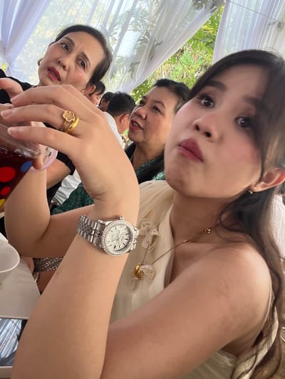 This close-up, low-angle photograph captures a social gathering in Bình An, Vietnam. The primary subject is a young woman in the foreground, looking directly at the camera with a slightly playful or pouty expression. She has long, dark hair and is wearing a light-colored garment, possibly off-shoulder, accessorized with a delicate gold-toned necklace. On her left wrist, she wears a silver-toned wristwatch featuring a diamond-studded bezel and a sparkling dial, while a gold-toned ring with a clover-like design adorns her right ring finger. She holds a transparent cup decorated with colorful circular patterns (red, blue, green).

Behind the main subject, two older women are partially visible. One woman, positioned in the mid-ground, appears to be smiling subtly. Another older woman, lower and to the left, has a more neutral expression and wears red lipstick. The head of a man is also partially visible between the figures.

The setting is an open-air or semi-outdoor event, suggested by the presence of white fabric draped overhead, resembling a tent or canopy. Lush green foliage is visible in the background, indicating a daytime event. White ceramic cups or bowls are also visible on a surface in the lower right, suggesting a meal or refreshments are being served. The image conveys a sense of intimate observation within a festive or communal atmosphere. There is no visible text.