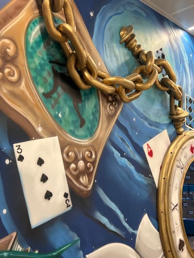 The image shows a wall mural inspired by Alice in Wonderland. A large, gold-colored chain dominates the foreground, looping across the frame. To the left of the chain is a golden, ornate frame with a teal-colored background, depicting a silhouetted figure with legs in mid-stride. Below this frame, a playing card, the three of spades, is positioned vertically. To the right of the chain, another playing card, the Ace of Hearts, is visible at an angle. A portion of a gold pocket watch face is also seen on the far right, with Roman numerals and playing card suit symbols. The background is a swirling blue and white pattern, suggesting a dreamlike or chaotic environment. This mural is located in Vaganovo, Russia. There are no people or interactions depicted in the image.