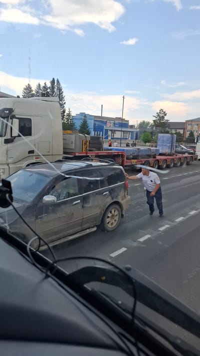 In Nizhny Ingash, Russia, a man in a white collared shirt and dark pants, likely a traffic controller or law enforcement officer, leans his right hand on the rear passenger door of a dark-colored SUV. The SUV is positioned on a road with white dashed lane markings. Behind the SUV, a long, flatbed trailer, laden with large blue cylindrical objects, is being towed by a white semi-truck. The trailer has multiple axles, indicating it's designed for heavy loads. In the background, a row of buildings lines the street, some painted in blue, with trees visible between them. The sky above is partly cloudy, with a mix of blue sky and white clouds. The overall scene appears to be an urban or semi-urban environment, possibly during daylight hours. The SUV appears dusty, suggesting it may have been driven on unpaved roads.