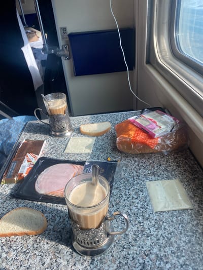 The image depicts the interior of a train compartment, situated in a train known to be in or passing through Kotelnich, Russia. A speckled gray and white tabletop, consistent with a fold-down train table, occupies the foreground. On the table, various food items are arranged: two glasses containing a light brown, foamy beverage, likely coffee with milk, are seated in ornate silver-colored metal podstakanniki (glass holders), each containing a spoon. Three separate slices of white bread are present on the table surface. A plastic-wrapped package of sliced pink luncheon meat or ham is visible, along with two individual, unwrapped slices of light-colored cheese. Additionally, a clear plastic bag contains a loaf of orange-colored bread, topped with a smaller plastic package of cheese slices. A brown rectangular package, possibly chocolate, with Russian text and an expiration date is also on the table. A white charging cable runs across the table, extending towards a window on the right side of the frame, which shows a bright, possibly sunlit exterior. To the left, a blue plaid blanket or fabric covers a surface. A partial view of an interior doorframe is visible in the background, displaying black numbers "52" and "51" vertically, with "76 77 78" below, and a dark blue panel. Behind the doorframe, a glimpse of an interior with a white-linened bed and headrest is apparent. No individuals are visible.