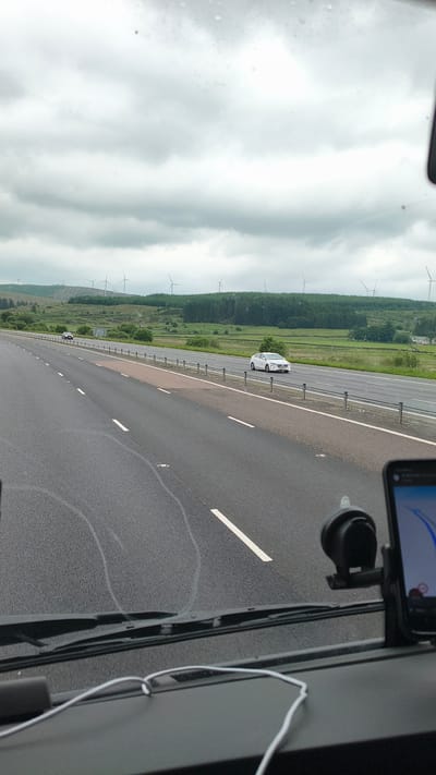 This is a landscape-oriented, eye-level, point-of-view shot from inside a vehicle, looking out at a highway. The scene is set on a cloudy day, possibly in the late morning or early afternoon, as indicated by the diffused light. The road, wet from recent rain, stretches ahead, marked by white dashed lines. To the left of the highway, a lush green landscape unfolds, featuring a dense forest bordering rolling fields. Wind turbines are visible scattered across the landscape, suggesting a rural or semi-rural location in the United Kingdom, specifically Elvanfoot. A white car is traveling on the highway ahead, and another dark-colored vehicle is further down the road. The windshield of the vehicle shows water droplets and streaks. A smartphone, mounted on a suction cup holder on the dashboard, displays a navigation app. The right side of the frame shows the interior of the vehicle, including a windshield wiper and a white cable. The overall impression is one of travel through a scenic, possibly breezy, environment.