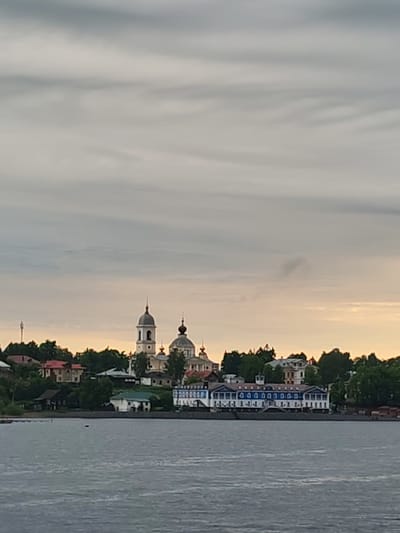The image depicts a view of Myshkin, Russia, from across the Volga River. The city skyline is visible, featuring the Dormition Cathedral (Uspenskiy Sobor) with its distinctive bell tower and onion domes. Various buildings and structures are nestled among trees along the riverbank. The river occupies the foreground, with its surface appearing slightly choppy. The sky is overcast, creating soft, diffused lighting across the scene.