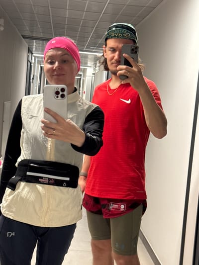 Two people, a man and a woman, are taking a mirror selfie. The man is wearing a red shirt with a white Nike logo, a patterned bandana, olive shorts, and a gold necklace. The woman is wearing a pink headband, a black long-sleeved shirt, a light beige vest, and dark pants. She has a black runner's belt around her waist. The background appears to be a brightly lit public restroom with tiled walls. It is unlikely that the location is Verkhnii Liuk, Russia, as the environment doesn't strongly indicate it.