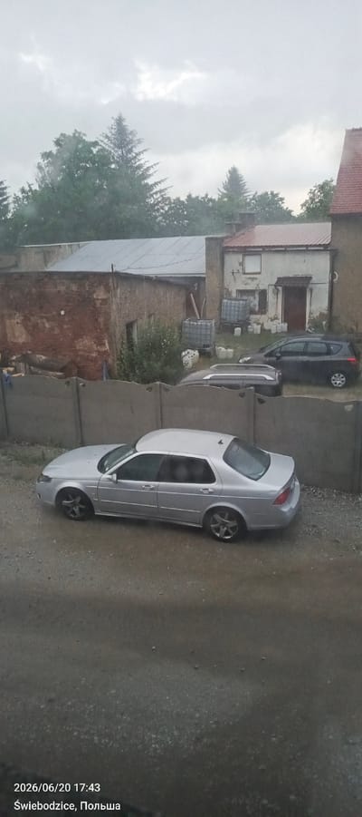 The image depicts an outdoor scene in Świebodzice, Poland, on June 20, 2026, at 17:43. A silver sedan is parked on a gravel surface in front of a concrete wall. Behind the wall, there are several buildings, including one with a reddish-brown brick facade and another with a white exterior and a red-tiled roof. The sky is overcast with visible clouds. Trees surround the buildings. There are also some industrial-looking containers and white plastic containers visible in the background.