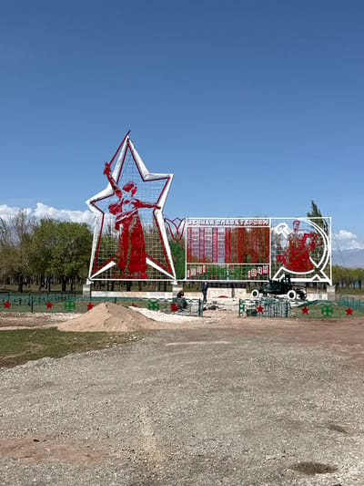 The image captures a memorial complex in Оттук, Kyrgyzstan, under a clear, bright blue sky during daylight hours. In the foreground and center-left, a prominent white five-pointed star, constructed from a metal framework with a grid pattern, encases a red silhouette of a female figure. This figure appears to be holding a wreath or torch with outstretched arms.
To the right and slightly behind this large star, another monument features a red silhouette of a Soviet soldier, equipped with a helmet and a weapon, positioned within a stylized circular or C-shaped emblem, possibly incorporating a hammer and sickle design. Below the soldier's monument, a rectangular section displays the Cyrillic text "ВЕЧНАЯ СЛАВА ГЕРОЯМ" (Eternal Glory to Heroes) alongside the dates "1941" and "1945," clearly indicating a dedication to the Great Patriotic War. Vertical panels on either side of the inscription likely serve as memorial plaques.
The monuments are situated on a grassy area, bordered by a green fence or railing adorned with small red five-pointed stars. A wide dirt or gravel path runs across the lower and right sections of the image. In the background, trees are visible under the sunny sky, with faint clouds in the distance. The scene is static, showcasing the architectural elements of the memorial.