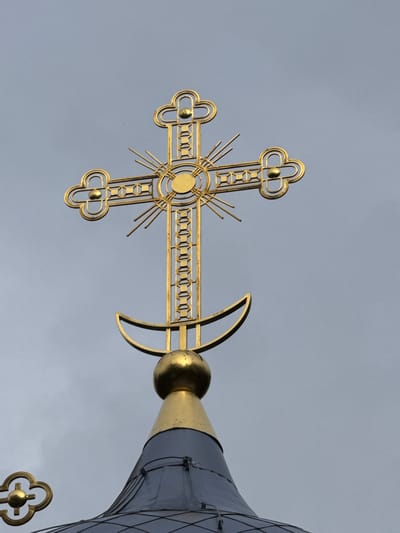 This is a close-up, low-angle shot of a golden Orthodox Christian cross atop a building, possibly a church or chapel, in Neniymaky, Russia. The cross is ornate, with a central circular element surrounded by radiating lines and decorative flourishes at its four ends. A small golden sphere is visible just to the right of the cross, attached to a golden conical structure that is part of the building's roof. This conical structure is dark gray and appears to be made of metal panels. The sky behind the cross is overcast and a uniform light gray, suggesting a cloudy day. The lighting is even, indicating it is likely daytime, though the cloud cover diffuses the sunlight. The image is well-focused on the cross and the adjacent architectural elements, with the background sky out of focus. The overall mood is solemn and serene, emphasizing the religious iconography and the grand scale of the structure.