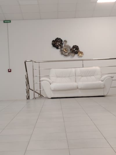 The image depicts an indoor scene, likely a public or commercial space. A white leather sofa is centrally placed against a white wall. Above the sofa, a metallic wall decoration resembling flowers is mounted. To the left of the sofa, a silver railing with vertical supports is visible, suggesting a staircase or a mezzanine. A red fire alarm is mounted on the wall below a green exit sign labeled "ВИХІД". The floor is tiled with light-colored square tiles. The lighting appears to be from overhead fluorescent fixtures. The overall impression is one of a modern, clean interior. There are no people visible in the image. The location is identified as being in the city of Chaiky, Ukraine.