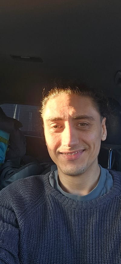 A man is pictured from the chest up, seated in what appears to be a car. He has his hair pulled back and is looking towards the camera with a slight smile. Sunlight is hitting the right side of his face, creating a strong shadow on the left. He is wearing a dark, textured sweater over a lighter t-shirt. In the background, to his left, are bags, one of which is a light blue backpack. The overall scene is dimly lit except for the sunlight on his face. There is no visible indication of the city of Neustadt-Glewe, Germany.