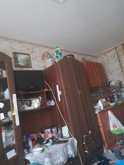 The image depicts a cluttered room, likely a living room or bedroom, in Shostka, Ukraine. The scene is dominated by wooden furniture, including a large, dark wood desk or cabinet that occupies the center of the frame. On this surface, various objects are scattered, such as a white plastic water dispenser, a black television, a framed picture of a landscape, and several smaller decorative items and boxes. To the left of the desk, a doll in a green and white outfit is visible, along with another doll or figurine. The wallpaper in the background features a floral pattern in muted tones of pink and beige.
Above the desk, a light-colored bag with a green logo is placed on top of some cabinetry. Further in the background, more wooden cabinets with doors are visible, along with a collection of household items. The overall impression is one of a lived-in, somewhat disorganized space. There is no clear indication of the time of day or weather conditions. The presence of the dolls and the general clutter suggest a domestic setting.
Visible text includes "Philips" on the television screen and part of a logo on the white bag. Some text is also visible on a card or paper placed on the lower shelf of the cabinet. The image is taken from a slightly elevated, tilted angle.