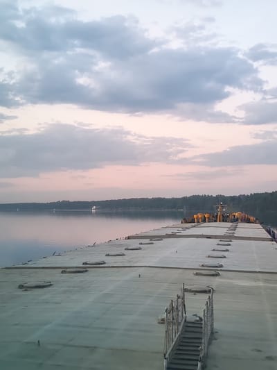 The image depicts the deck of a barge or ship on a body of water, possibly the Protva River near Ermolino, Russia, given the provided location. The deck is flat, light gray, and has several circular hatches or access points. A metal staircase leads down to the deck from the foreground. The water is calm and reflects the light of the sky. A line of trees is visible on the distant shore. The sky is cloudy with hues of gray, white, and pink, suggesting either sunrise or sunset. The lighting is soft and diffused.