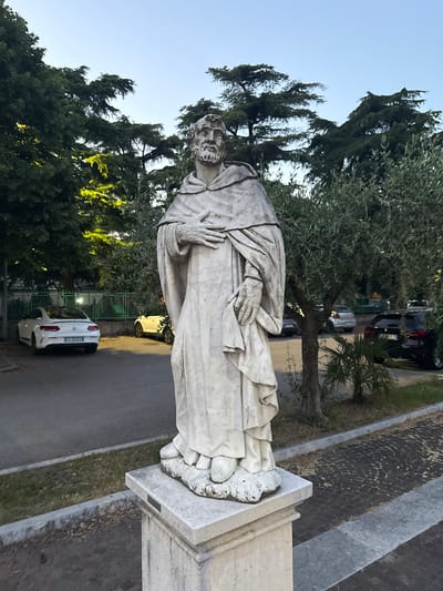 The image shows a marble statue of a bearded man, possibly a saint or historical figure, positioned on a stone pedestal. The statue is draped in flowing robes. The setting appears to be an urban environment in Villafranca di Verona, Italy. In the background, there are trees, a street with cars parked on it, and a glimpse of other buildings. The sky is visible in the upper portion of the image. The statue is centrally located and appears to be a permanent fixture.