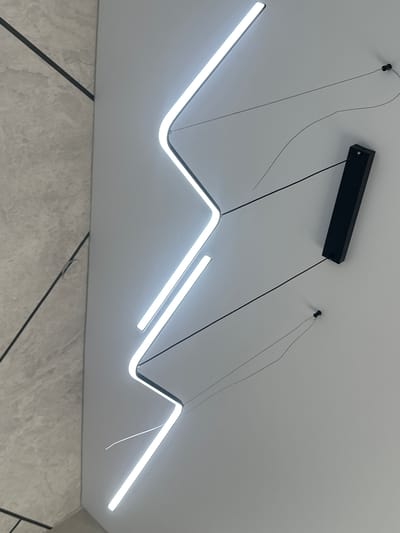 The image shows a modern interior with a white wall and a grey, textured wall on the left. A zigzagging LED light fixture is mounted on the white wall. A black rectangular electrical junction box is also visible on the white wall. The scene is illuminated by the LED light. The location in the city of Tabuny, Russia is not discernible from the image.