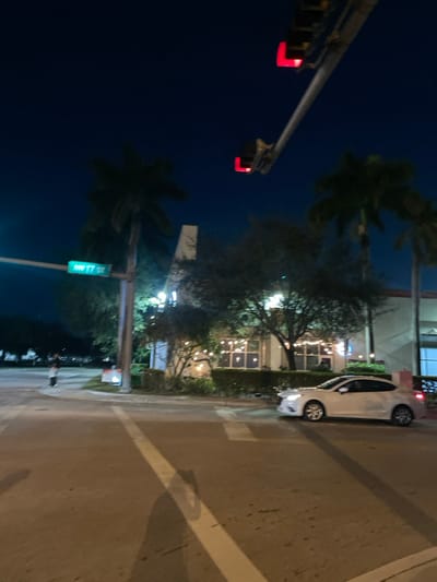 A nighttime scene depicts a white car parked on a street next to a building with lit windows. Palm trees are visible on both sides of the street. A traffic light structure is present with red lights illuminated. A green street sign reading "NW 17 St" is also visible on a pole. The street surface has white lines painted on it.