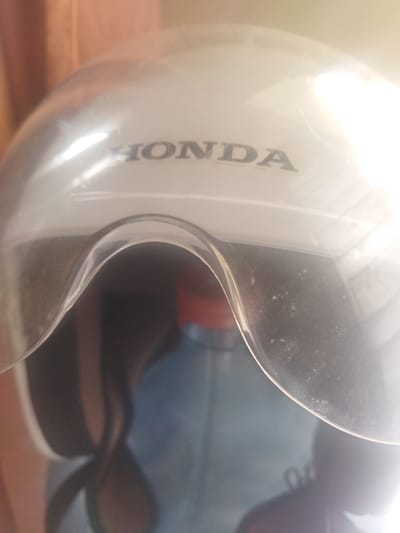 This is a close-up, slightly blurry image of a white motorcycle helmet with a clear visor. The "HONDA" logo is visible in dark grey lettering on the top surface of the helmet. The visor curves downwards in front of the helmet, obscuring the lower part of the helmet's interior, where a red and blue object is partially visible. The background is out of focus, with vertical lines suggesting fabric or blinds, in muted brown and orange tones. Light reflects strongly off the helmet's surface, particularly on the right side of the image. The overall impression is of a product shot or an indoor scene with artificial lighting. There is no clear indication of time of day, weather, or specific emotions. The text "HONDA" is the only discernible text in the image. The location context provided is Lancokpanteara, Indonesia, but this information is not visually evident in the image itself.