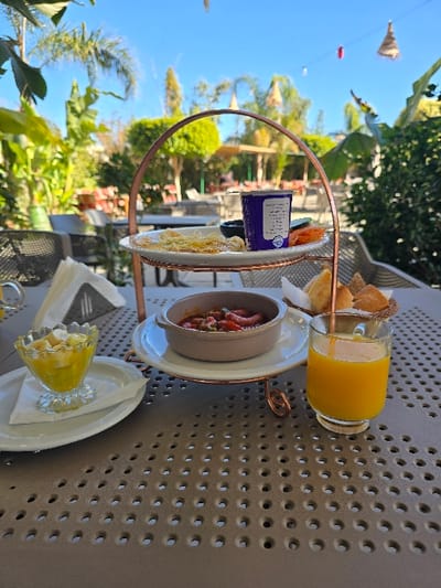 The image depicts an outdoor dining setup on a clear, sunny day at a restaurant or cafe in Taza, Morocco. A grey perforated metal table occupies the immediate foreground. On the table, from left to right: a clear glass bowl containing a fruit salad with yellow and white fruit pieces, resting on a small white plate with a folded white napkin; a two-tiered, copper-colored metal serving stand; a clear glass filled with orange juice; and a small wicker basket holding several bread rolls.

The lower tier of the serving stand supports a light brown ceramic bowl with handles, containing a red stew-like dish with visible sausage segments and vegetables, placed on a white plate. The upper tier holds a white plate displaying several thin, yellowish food items (resembling crepes or pancakes), a small dark bowl (contents indistinct), several small orange slices, and a tall blue cup with white labeling and an inserted stirring utensil.

The background reveals a vibrant outdoor garden or patio environment, characterized by abundant green foliage, including palm trees, under a bright blue sky. Multiple unoccupied outdoor tables and chairs, appearing to be dark-colored, are arranged in the mid-ground. Overhead, decorative elements are suspended, including red lanterns and light-colored woven conical shapes. No individuals are present within the frame.