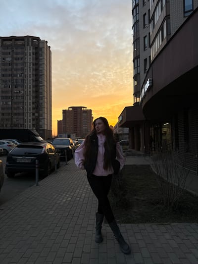 The image displays an urban street scene in Gribovo, Russia, illuminated by the warm, low light of either sunrise or sunset. A young adult female stands on a gray paved sidewalk in the foreground, facing slightly left of the camera with her gaze directed to the left. She has long, dark hair and is wearing a light pink hooded sweatshirt, a dark vest, dark leggings, and dark, chunky boots; her hands are in her vest pockets. To her left, a row of parked vehicles lines a dark asphalt street, including a black Ford Focus sedan with the Russian license plate O267EO 197. Behind the vehicles, two tall, multi-story residential buildings with numerous windows and balconies are visible. To the female's right, a modern commercial building with a dark facade and a curved section features a prominent, glowing white sign displaying "КОФЕ" (Coffee). A narrow strip of bare ground with sparse grass and leafless shrubs separates the sidewalk from this building. The sky exhibits orange and yellow hues with scattered clouds, backlighting the scene.