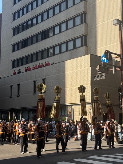 This daytime image depicts a street scene during a festival parade in Kanazawa, Japan, likely the Hyakumangoku Matsuri. The perspective is slightly elevated, looking down a city street towards a multi-story building.
In the foreground and mid-ground, a procession of approximately 20-30 individuals, predominantly men, advances on a paved street. They are dressed in traditional festival attire consisting of black trousers, short-sleeved jackets (some brown with black stripes, others patterned), and white headbands. Several men carry long wooden poles. Prominently featured are five tall, decorative elements, each topped with a gold, five-petaled motif displaying kanji characters. From left to right, these read "松ヶ枝" (Matsugae), "祭山" (Matsuyama), "祭山" (Matsuyama), "戸板" (Toita), and "松ヶ枝" (Matsugae). Beneath these gold tops are cylindrical structures composed of vertical red and gold, or gold and black, tassels or strips. The individuals holding these elements typically have them raised above their heads or shoulders. Spectators line the sides of the street behind the procession. The street surface includes a visible white crosswalk in the lower right.
In the mid-ground, on a lower-level balcony or terrace of the multi-story building, a group of approximately 10-15 women is observing the parade. They are attired in red kimonos, some with dark patterns, and have traditional hairstyles.
The background is dominated by the multi-story building, which features a light-colored brick or tiled facade and multiple rows of rectangular windows with dark frames. The building exhibits recessed sections on some floors, creating architectural variation. On the right side of the image, a dark metal pole supports a set of three dark-lensed traffic lights and a circular blue sign with a white left-turn arrow. A square-shaped street light fixture is also mounted on the pole. Strong sunlight illuminates the upper right portion of the building, casting distinct shadows across the street and lower parts of the building.