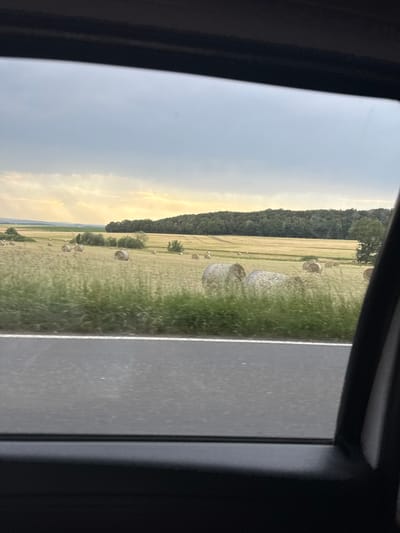 This is a photograph taken from inside a vehicle, looking out the window at a rural landscape. In the foreground, a grey asphalt road with a white line marking its edge is visible. Beyond the road, a field of dry, yellow grass and some green vegetation stretches out. Several round hay bales are scattered across this field. In the middle ground, a dense line of dark green trees forms a treeline. Further in the background, rolling hills are visible under a cloudy, grey sky with some patches of yellow light suggesting the sun is breaking through. The scene appears to be located in the vicinity of Oberwetz, Germany, based on the context provided. There are no people or discernible animals in the image. The overall impression is that of a countryside vista captured during a drive.