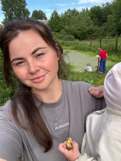 The image shows a woman in the foreground, holding a piece of food and looking at the camera with a slight smile. She is wearing a grey t-shirt with a Kappa logo. Behind her, a pond with green water is visible, with two children and an adult man in the background. The man is wearing a red shirt and blue pants, and he appears to be interacting with a child near the pond. The environment is rural, with trees and vegetation surrounding the pond. The overall scene suggests a casual outdoor gathering in a village setting, possibly in Малая Рукавицкая, Russia, given the context provided.