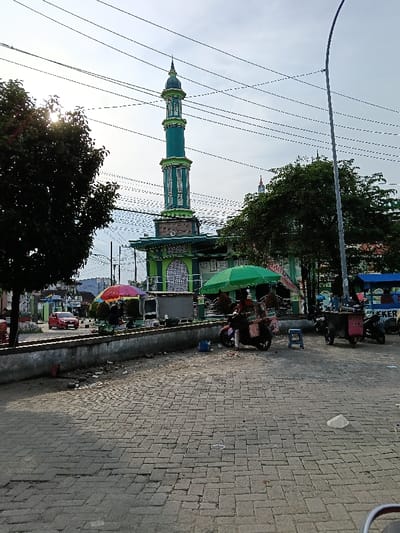 The image depicts a daytime street scene in Sumberejo, Indonesia, centered around a green and white mosque with a tall, distinctive minaret. The minaret is prominently featured against a bright, slightly overcast sky, with numerous overhead power lines crisscrossing the frame.

In the mid-ground, a low concrete wall separates a paved area of interlocking bricks from the elevated ground surrounding the mosque. Along this wall and near the mosque's perimeter, several street vendor operations are visible. On the left, a stall with a multi-colored (red, orange, yellow) umbrella is set up. Closer to the mosque, under a green umbrella, is a vendor on a dark-colored scooter with a merchandise box attached to its rear. A small blue plastic stool sits nearby. Further to the right, another mobile vendor cart, partially obscured, shows the label "LEKER" and has a blue canopy. Multiple motorcycles are parked near these vendor stalls. Indistinct human figures are present near the stalls, suggesting commercial activity.

To the left, beyond the concrete wall, a red car is parked on a street, with other buildings visible in the background. A dark green tree on the far left partially obscures the sun, creating a visible lens flare. Another dark green tree is situated to the right of the mosque. A tall, dark street light pole stands on the far right of the image. The paved foreground shows minor debris.