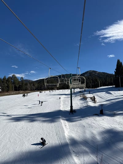 A ski lift with empty chairs ascends a snowy slope under a clear blue sky with wispy clouds. Sparse trees line the edge of the ski run, where skiers and snowboarders dot the white expanse. Shadows cast by the lift's support structures stretch across the snow. The scene conveys a sunny day at a ski resort with a relaxed, open atmosphere.
