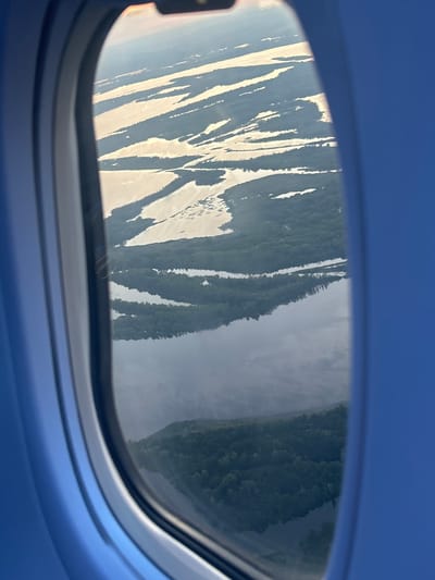 An aerial view through an airplane window shows a vast, sun-drenched river delta near Kokkorevo, Russia. The delta is characterized by numerous islands and channels of water reflecting the sunlight, creating bright, irregular shapes against the darker green of the surrounding forested land. The foreground is dominated by the blue interior of the airplane cabin, framing the landscape. The water appears calm, with the sun casting a warm glow on its surface. The dense green foliage indicates a healthy ecosystem with abundant trees. The overall impression is one of serene natural beauty viewed from a great height.