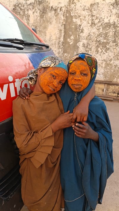Two young individuals, likely girls, are captured in a portrait-style photograph, standing close together in an affectionate embrace. They are positioned in front of a red and blue vehicle, possibly a van or truck, which has white lettering on its side, partially revealing "vito" or "vita". The background behind them is a light-colored, textured wall, possibly stucco or concrete, showing signs of wear and age.

Both individuals have their faces, ears, and parts of their neck covered in a striking, coarse, orange substance, possibly a ceremonial paste or pigment. This substance gives their skin a distinct, vibrant hue and textured appearance.

The individual on the left wears a light brown, flowing garment, possibly a modest traditional dress or abaya, with distinctive layered or pleated sleeves. She also wears a patterned head covering, predominantly blue and white with black details, which frames her face. Her right arm is wrapped around the shoulder of the person on the right, and her left hand is clasped with the other person's left hand. She looks directly at the camera with a soft, gentle smile.

The individual on the right wears a dark teal or dark blue, flowing garment, also likely a traditional dress, with a visible zipper down the front. Her head is covered by a colorful patterned fabric featuring shades of blue, yellow, orange, and black. Her left arm is wrapped around the waist or side of the person on the left, and her left hand is clasped with the other person's left hand. Her right hand is visible, resting near her lower torso. She also looks at the camera with a gentle, slightly smiling expression.

The overall impression is one of companionship and cultural significance, highlighted by the unique facial decoration. The setting appears to be outdoors during daylight, with diffused lighting, suggesting either an overcast day or shade.