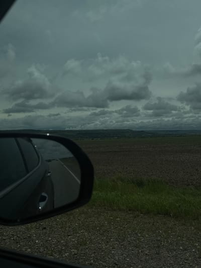 The image depicts a view from a car window, possibly while driving. A side mirror is visible, reflecting a portion of the road and the car itself. Outside the car, a landscape of fields stretches towards the horizon under a cloudy sky. The scene might be from Svetlograd, Russia, given the location information. The road surface appears to be asphalt, bordered by a shoulder of gravel and grass. No people are visible in the image.