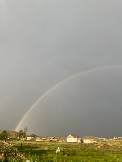 A wide-angle shot captures a rural landscape with a prominent rainbow arcing across a cloudy sky. The scene is set in the city of Kara-Oi, Kyrgyzstan. In the foreground, lush green grass covers a gently rolling terrain. A cluster of modest, weathered buildings, including houses and what appears to be a small farm or village, are visible in the middle ground. The architecture suggests a traditional or rural style, with simple structures and fenced-off areas. The sky is overcast, with dark, gray clouds dominating the upper portion of the frame, creating a dramatic backdrop for the vibrant rainbow. The overall impression is one of natural beauty and a tranquil, albeit somewhat rustic, rural environment.