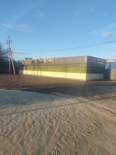 In the city of Aral, Kazakhstan, an exterior view captures a cafe with a corrugated metal roof and siding. The lower section of the building is adorned with a textured green material, possibly artificial turf, and a white patterned facade. The name "HYP КАФЕ" is prominently displayed in large, stylized letters on the upper part of the building, with additional text in what appears to be Cyrillic script to the right.
In front of the cafe is a lot covered with dark gravel and dirt, showing clear tire tracks from vehicles. A light pole stands to the left of the building, with wires extending from it. The sky above is a clear blue with a few wisps of clouds, and the sun casts shadows on the ground, indicating it's either early morning or late afternoon. The overall scene depicts a functional establishment in a somewhat unpaved area, typical of some parts of Kazakhstan. There are no people visible in the image.