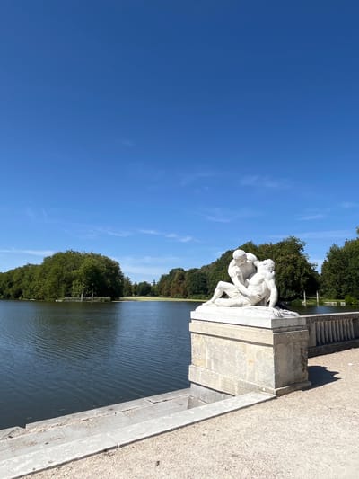 The image depicts a serene outdoor scene with a classical marble sculpture positioned in the foreground. The sculpture, situated on a stone pedestal, features two male figures in a dynamic embrace, rendered in white marble. Behind the sculpture, a tranquil body of water stretches out, bordered by lush green trees and a clear blue sky with scattered wispy clouds. The environment appears to be a park or garden, possibly part of the Château de Rambouillet in Rambouillet, France, given the style of the sculpture and the surrounding landscape. The sunlight casts shadows on the ground, indicating a bright, sunny day.