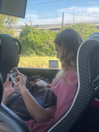 The image captures two individuals inside a vehicle, identified as being in Linden, United States. A male subject with dark, curly hair and a beard, wearing a dark grey t-shirt, is reclined with his head resting on the lap of a female subject. He is actively engaged with a white-cased smartphone, holding it with both hands and viewing its screen. The phone case features several small photographic images on its back. A light-colored bracelet is visible on his left wrist. The female subject, positioned to the right, has long, light brown hair and is wearing a pink long-sleeved top. Her gaze is directed downwards towards the male or the smartphone. The vehicle's interior features dark grey seats, and an "EMERGENCY EXIT" sign with a right-pointing arrow is visible through the window. The exterior environment consists of green foliage and dry grass in the foreground, with a concrete overpass, utility poles, and overhead wires visible against a bright blue sky with sparse clouds in the distance.