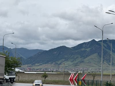 The image presents an overcast daytime scene of a road traversing a mountainous landscape near Korkmaskala, Russia. In the immediate foreground, a multi-lane asphalt road occupies the bottom of the frame, with two vehicles visible. On the left, part of a large white truck is seen, with Cyrillic text "ПРОФИЛЯ" (Profilia) visible on its side. On the right, a silver sedan with its headlights illuminated drives towards the viewer. The car's license plate displays "х 065 хс 05".

Numerous streetlights with curved poles extend over the road, and on the right, distinctive red and white chevron signs indicate a curve or hazard. A low, light-colored brick wall and a green metal fence separate the road from the midground. Beyond this barrier, a small town or village is nestled in the valley, featuring a cluster of houses, some with colorful roofs, and a more prominent, multi-story pinkish building. Sparse green trees are scattered along the roadside and among the buildings.

The background is dominated by large, rugged mountains covered in green vegetation, indicating an area with significant natural terrain. The sky above is entirely covered by thick, grey clouds, suggesting an overcast day with subdued light. The overall scene depicts a functional road leading through a populated valley surrounded by the formidable natural beauty of mountains, under a calm, cloudy sky.