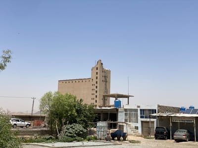 The scene is a wide shot of a building complex in Panjwai, Afghanistan (latitude: 31.615, longitude: 65.589). It is a sunny day with a clear blue sky. The main structure is a large, tan, rectangular building with several windows and an apparent flat roof. The building is located in a rural area with a few other smaller structures around it. There are a couple of cars parked in front of the building, one of which is white and the other is dark gray. There are several trees and shrubs in front of the building, along with a few other miscellaneous items. The building appears to be in a state of disrepair, with some of the windows broken and the paint chipped. The smaller structures also appear to be in disrepair. There are some power lines running across the scene.