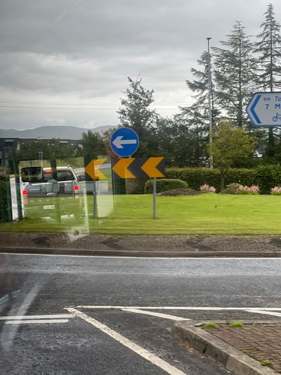 The image shows a view from a vehicle, likely looking out the side window. The scene is a street with a circular grassy area, possibly a roundabout, bordered by hedges and trees. Road signs are prominent: a blue circular sign with a white arrow pointing left, and yellow chevron signs indicating a sharp left turn. In the background, there are mountains, a cloudy sky, and some buildings with solar panels. A silver pickup truck is visible on the road, reflecting in the window. The road in the foreground is wet, suggesting recent rain. The overall impression is a roadside view in a somewhat overcast, possibly rural or suburban, setting in Ireland.