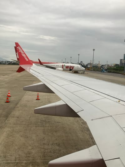 An overhead view captures a section of an airport tarmac at Da Nang International Airport, located in the vicinity of Hòa Cường Ward, Da Nang, Vietnam, under an overcast sky. In the immediate foreground, the clean white wing of a passenger aircraft, featuring a red wingtip and visible flaps, extends from the lower right towards the center-left. Three orange and white traffic cones are positioned on the concrete tarmac to the left of the foreground wing. In the midground, two parked commercial aircraft are visible: a red and white Vietjet Air aircraft, identified by "VietjetAir.com" on its tail, is positioned to the left. To its right, a white T'way Air aircraft, marked with "t'way" in red on its fuselage, is also parked. Several ground crew personnel are observable near the nose landing gear of the T'way Air aircraft, engaged in ground operations. Further in the background, tall light poles, indistinct buildings, and a mobile passenger staircase are present. A distant mountain range is faintly visible on the left horizon.