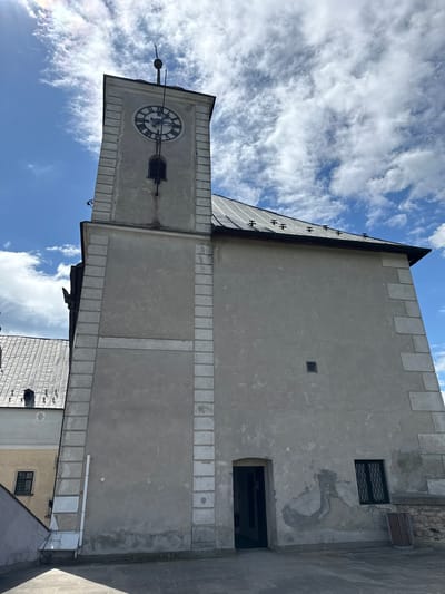 The image shows a clock tower in Častá, Slovakia. The tower is made of stone and has a clock face near the top. The sky is blue with white clouds. The tower appears to be attached to a building with a castellated roof. Other buildings are visible behind the tower.