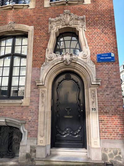 The image displays a detailed view of a building facade, specifically the entrance and adjacent architectural elements, located at Rue Paul Lauters 32 in Ixelles-Elsene, Belgium. The facade is constructed primarily of red brickwork. The central feature is a glossy black, ornately carved wooden door with curvilinear panels, an elaborate dark-colored door knocker, and a rectangular mail slot. The door is set within a highly decorative, light-colored stone archway featuring rococo-style carvings, including volutes and shell motifs. To the right of the archway, the number "32" is inscribed in black on the stone. Two shallow stone steps lead up to the door.

Above the entrance, an irregularly shaped window with black gridded glazing is framed by an equally elaborate sculpted stone surround. To the left of the entrance, a large rectangular window with black gridded glazing is set within a simpler, yet still ornate, stone frame. Below this left window, a small, recessed ground-level opening is secured by an intricate, dark metal grille with organic, swirling patterns. The building's base comprises a mix of cut stone blocks and rubble masonry. A blue street sign, partially visible on the brick wall to the right of the upper window, identifies the location as "IXELLES-ELSENE" and "RUE PAUL LAUTERS / STRAAT." The scene is illuminated by natural daylight, and reflections of a light-colored structure are visible in the windowpanes. No individuals or active subjects are present.