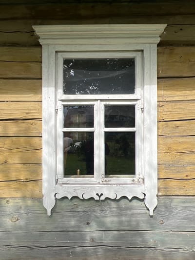 A weathered, white-painted wooden window is centered on a log wall of a traditional rural Belarusian house. The upper portion of the wall is constructed of horizontally laid, yellow-tinted logs with visible grain and occasional cracks. The lower portion of the wall is made of wider, grey-toned wooden planks, also showing natural wear and grain patterns. The window itself is divided into four panes, two on top and two on the bottom, framed by white-painted wood. The window frame features decorative elements, including a carved, scalloped trim piece below the lower panes and ornate finials on the sides. Reflections in the glass suggest an outdoor environment with trees and possibly a building visible. The scene appears to be in Бучацінскі сельскі Савет, Belarus, indicative of traditional rural architecture.