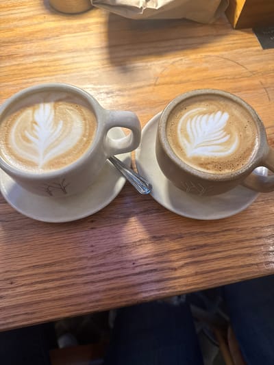 Two mugs of latte art coffee sit on a wooden table. One mug is tan and the other is light gray. Both mugs sit on matching saucers and the light gray mug has a spoon in it. A partially visible paper bag sits to the left of the mugs. A person is partially visible to the right. The location is San Cristóbal, Mexico.