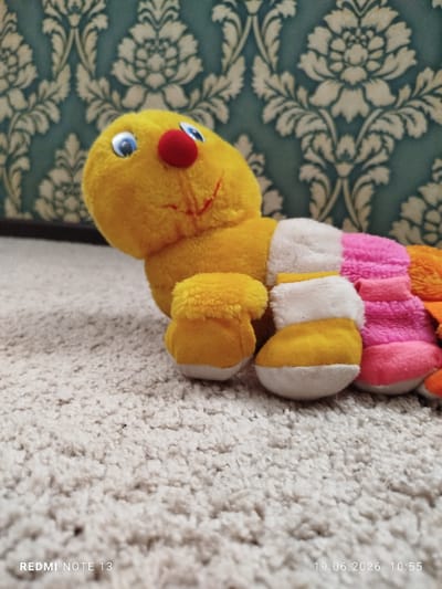 A plush caterpillar toy with a yellow head and body segments in white, pink, and orange lies on a cream-colored, textured carpet. The toy has blue eyes, a red nose, and a stitched red smile. The background features a teal wallpaper with a white floral damask pattern. The timestamp "19.06.2026 10:55" and "REDMI NOTE 13" are overlaid on the bottom of the image. The scene is indoors, with no indication of the specific location within the city of Асан-Чек, Kyrgyzstan.
