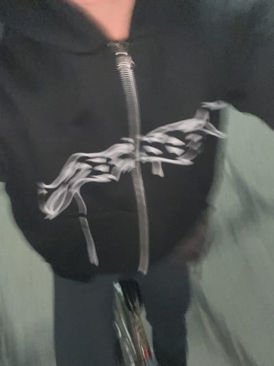 A blurry image shows a black garment, possibly a jacket or sweater, with a white graphic design. The design appears stylized, perhaps a skull or abstract figure, with sharp, angular features. A metal zipper runs across the garment. The background is a mix of light and dark tones. Given the blur, it's impossible to pinpoint the exact location in Olivos, Argentina.