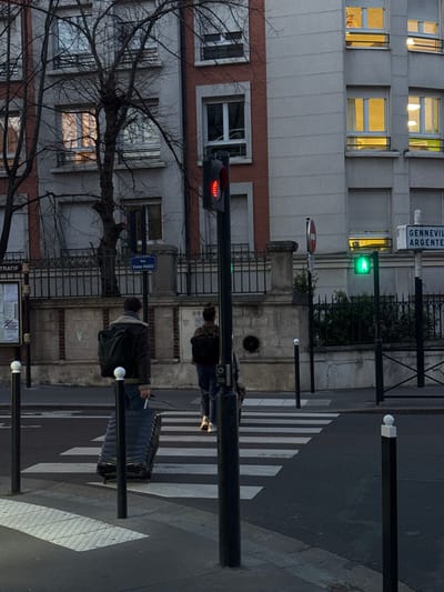 The image depicts an urban street scene at dusk or early evening on Rue Victor Hugo in Bois-Colombes, France. Two individuals are observed walking across a pedestrian crosswalk. The person in the foreground, viewed from behind, wears a dark jacket, a dark backpack, and is pulling a dark, ribbed, wheeled suitcase. A second individual, also viewed from behind, wears a dark top, dark pants, and a dark backpack, walking further along the crosswalk to the right.
In the mid-ground, a black traffic light pole stands centrally, displaying a red light for vehicular traffic. To its right, a pedestrian signal displays a green "walk" symbol. Street signs are visible; one identifies the location as "Rue Victor HUGO BOIS-COLOMBES," and another points towards "GENNEVILLES ARGENTEUIL." A red circular "No Entry" sign is also present. The sidewalk is separated from multi-story buildings by a black metal fence and bare-branched trees.
The buildings in the background consist of a structure on the left with red brick and gray sections, and another on the right primarily gray. Many windows in both buildings exhibit warm yellow interior lighting, with some windows on the right building also showing closed roller shutters. The ground is asphalt, and several dark bollards with white tops are positioned along the curb and street.
